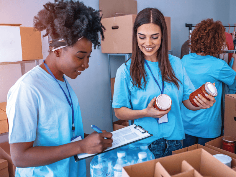 Two women in blue shirts sorting food donations in a warehouse during a volunteer food drive.