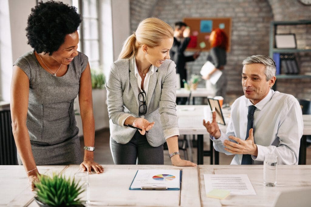 Group of smiling business people analyzing reports and communicating while working together in the office. There are people in the background.