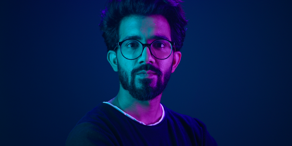 A man with a beard and glasses stands in front of a blue light, deep in thought.