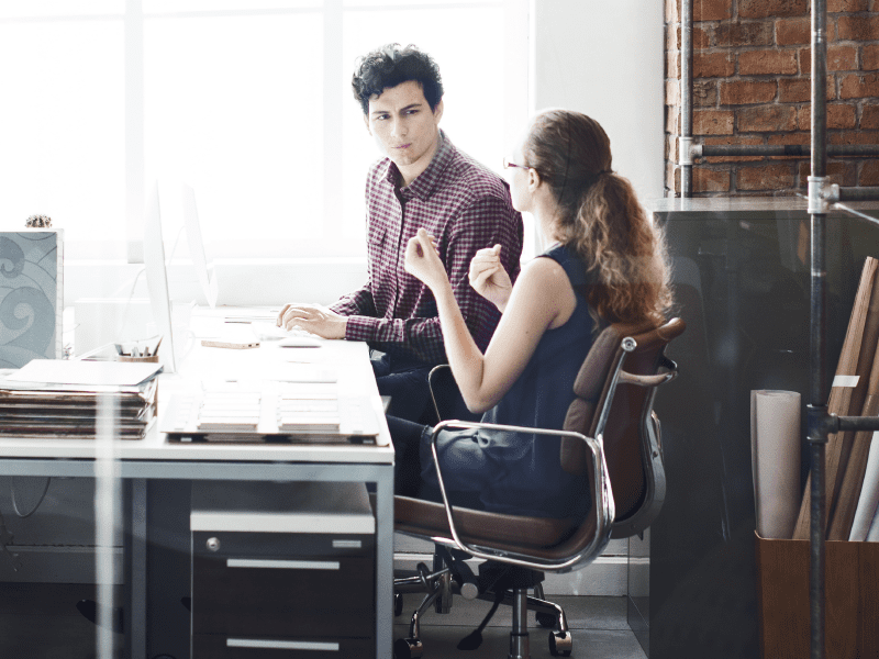 A man and woman sitting at a desk in an office, engaged in a professional discussion.