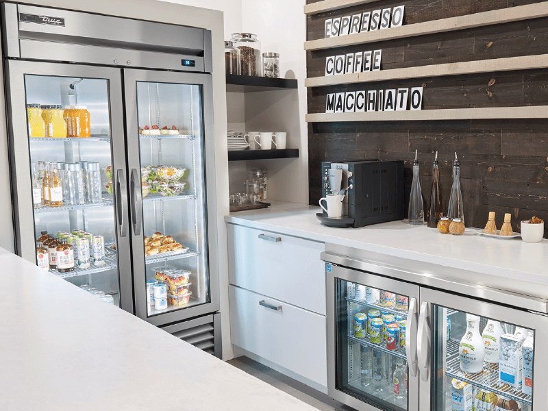 Modern cafeteria corner with fully stocked beverage fridges, a coffee machine, and a wooden sign listing coffee types above.