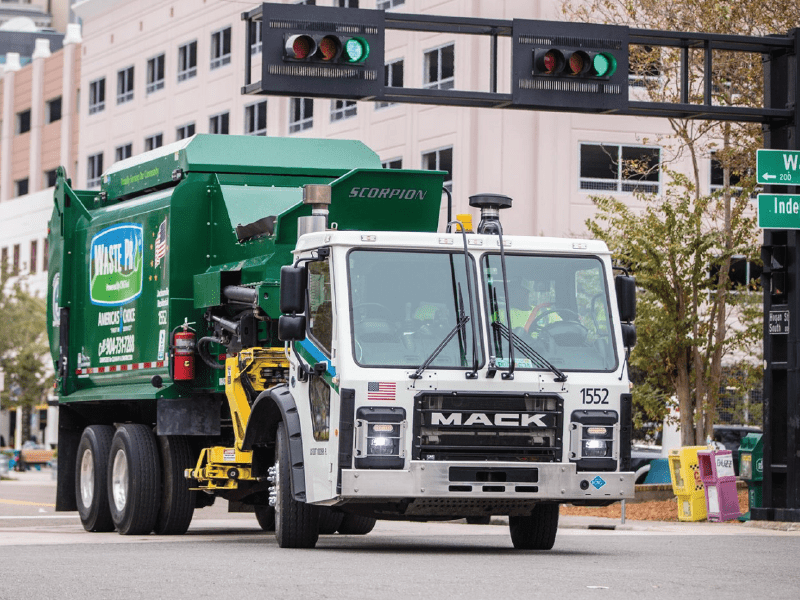 A green and white Waste Pro garbage truck on a city street, collecting trash with traffic lights showing green in the background.