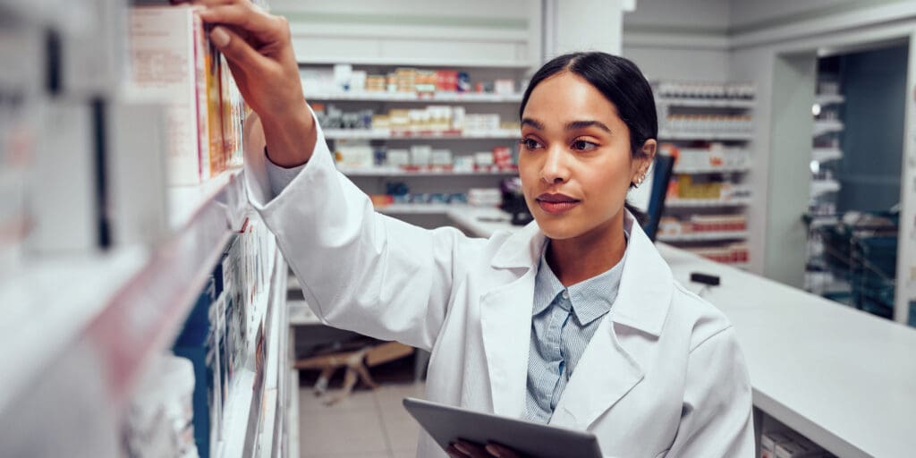 Woman Wearing Lab coat Holding Digital Tablet Searching for Medicine on Shelves of Pharmacy