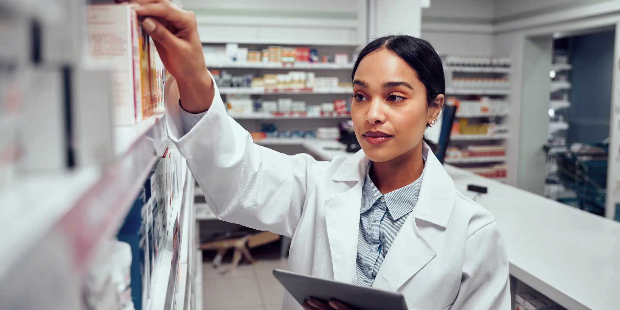 Woman Wearing Lab coat Holding Digital Tablet Searching for Medicine on Shelves of Pharmacy