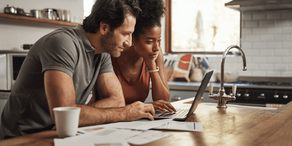 A couple is intently viewing a laptop in their kitchen, highlighting a moment of teamwork and shared interest in technology.