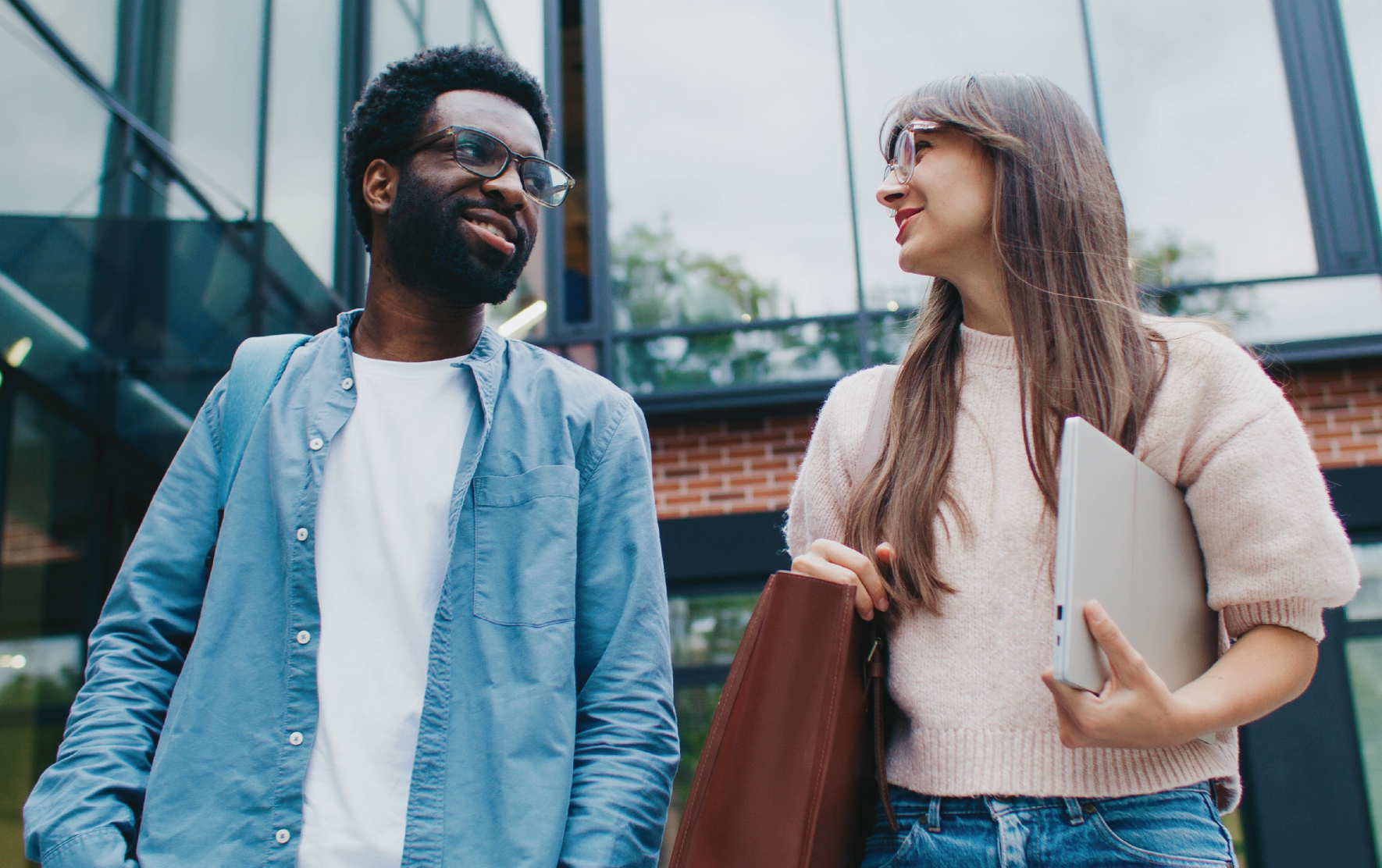 Two professionals walking outside a modern office building, carrying work essentials and appearing engaged in conversation.